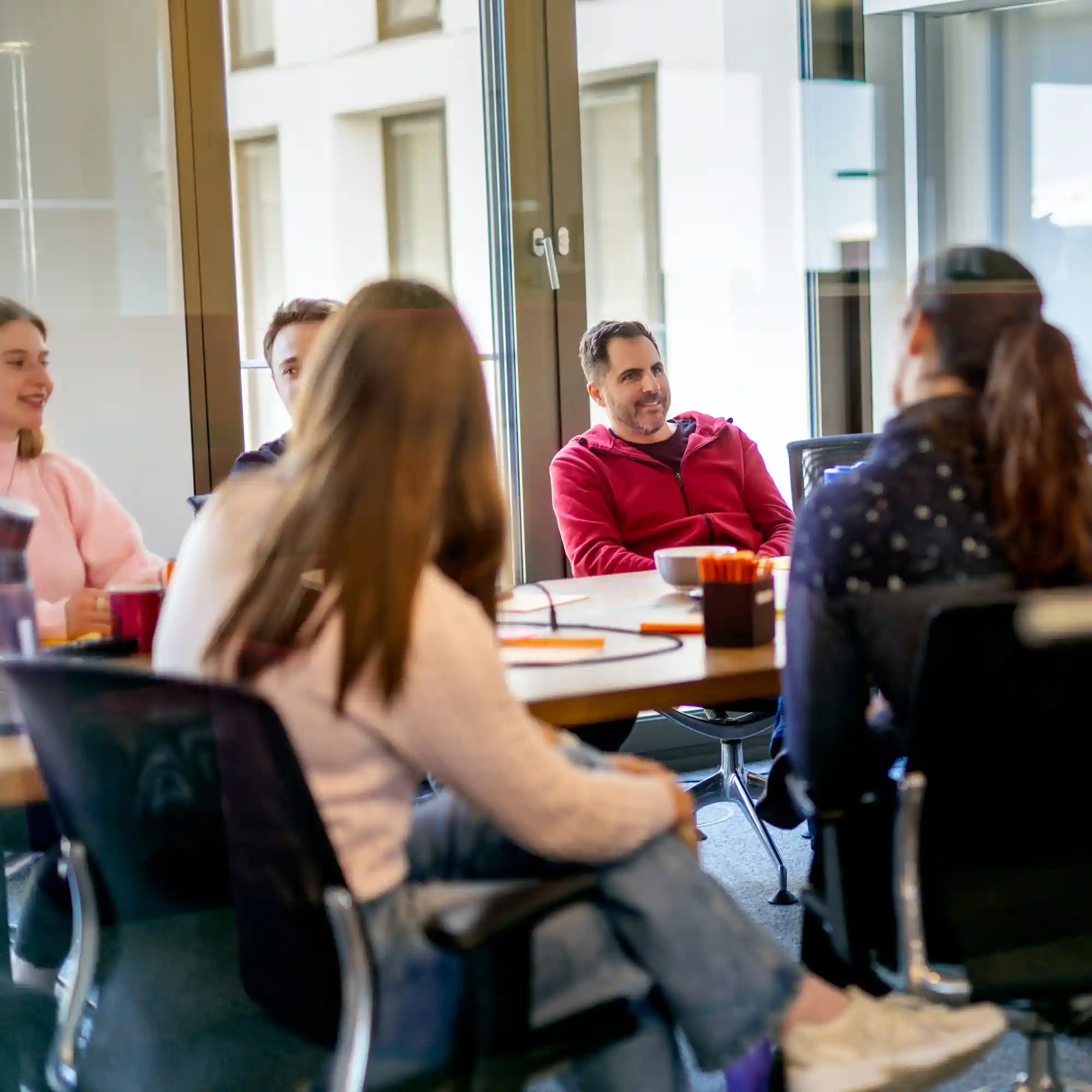 Team sitzt gemeinsam an einem Tisch in einem hellen Raum mit großen Fenstern. Auf dem Tisch liegen Notizbücher, Stifte und Tassen – die Szene zeigt eine Besprechung oder Zusammenarbeit.