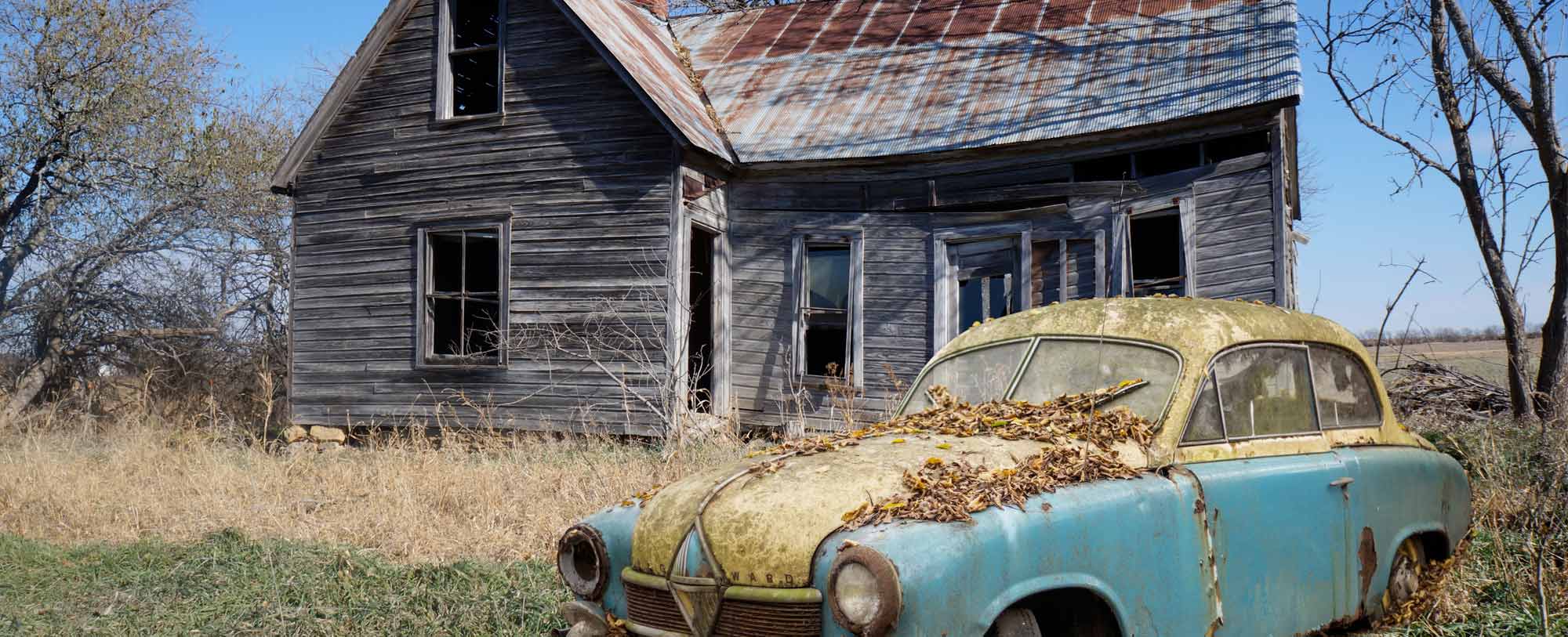 Rusted old car in the grass in front of a dilapidated house