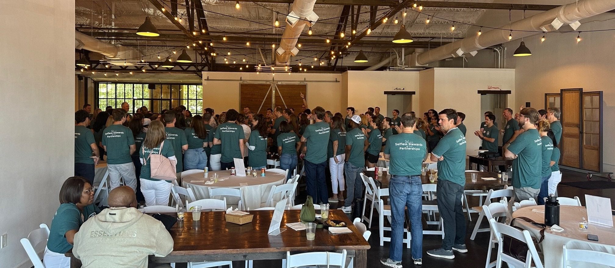 This picture highlights a corporate event at the Westside Warehouse's modern industrial venue space. String lights hand from the rafters and provide a warm glow to the space. The image features many guests participating in a group activity. They are all wearing the same green shirt, representing the company they work for. A mix of round tables and farm tables can be seen, as well as some decorative items.