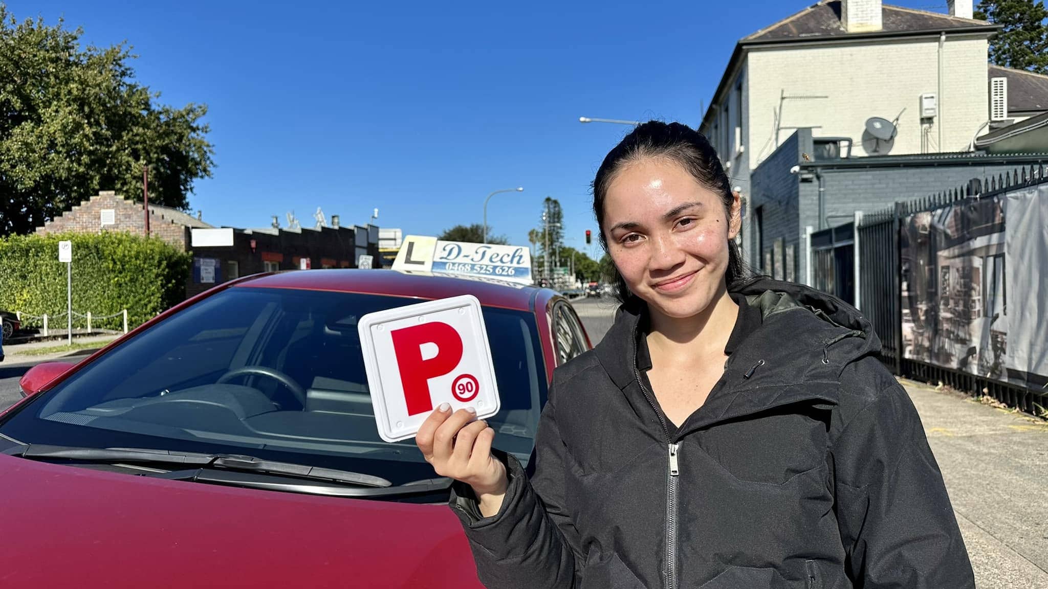 Smiling driving school student holding a red P-plate next to a D-Tech dual-control car after passing her practical driving test in Marsden Park.