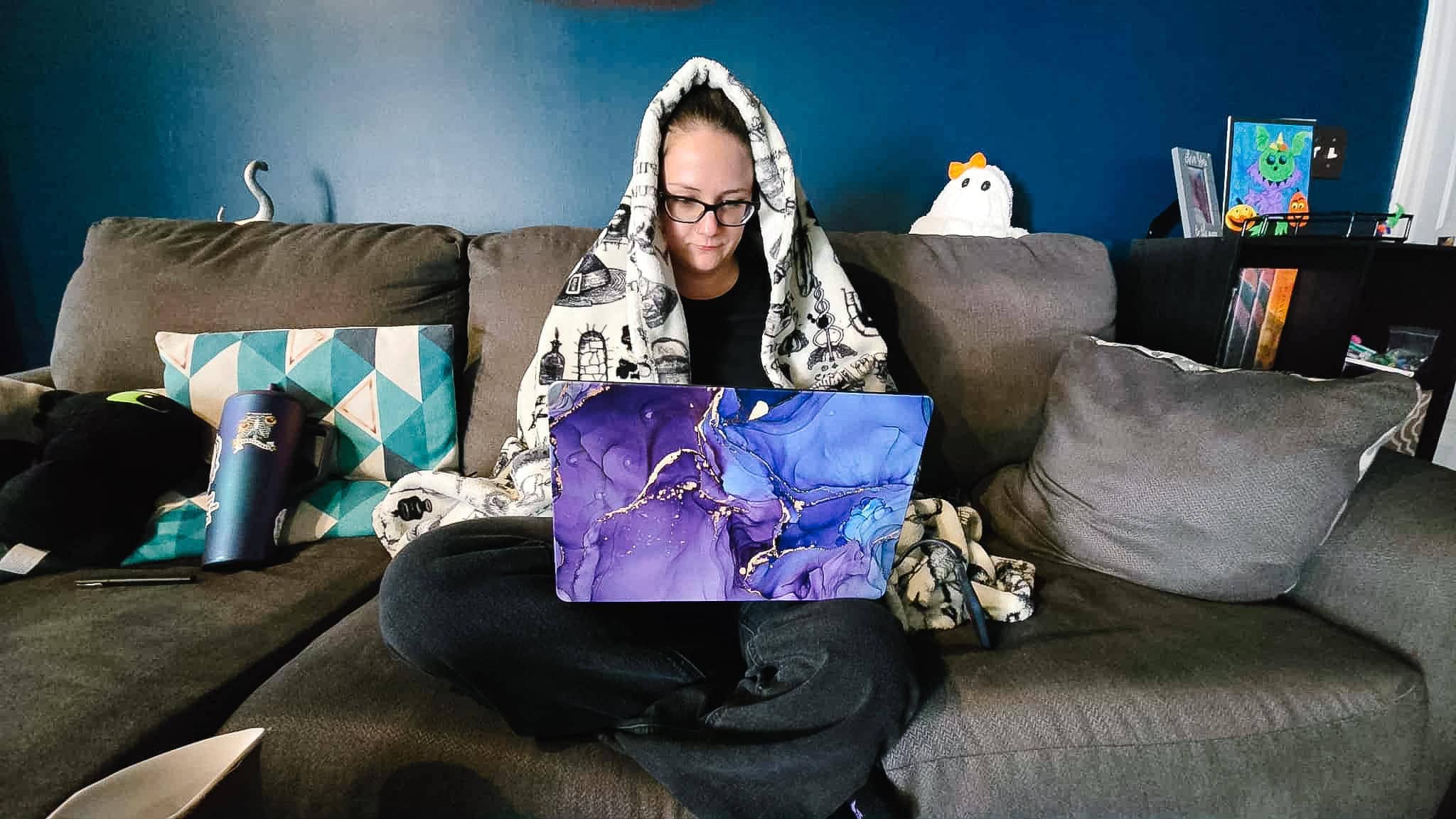 Phoenyx bundled in a blanket on the couch with a laptop, recovering from an autoimmune flare up after travel.