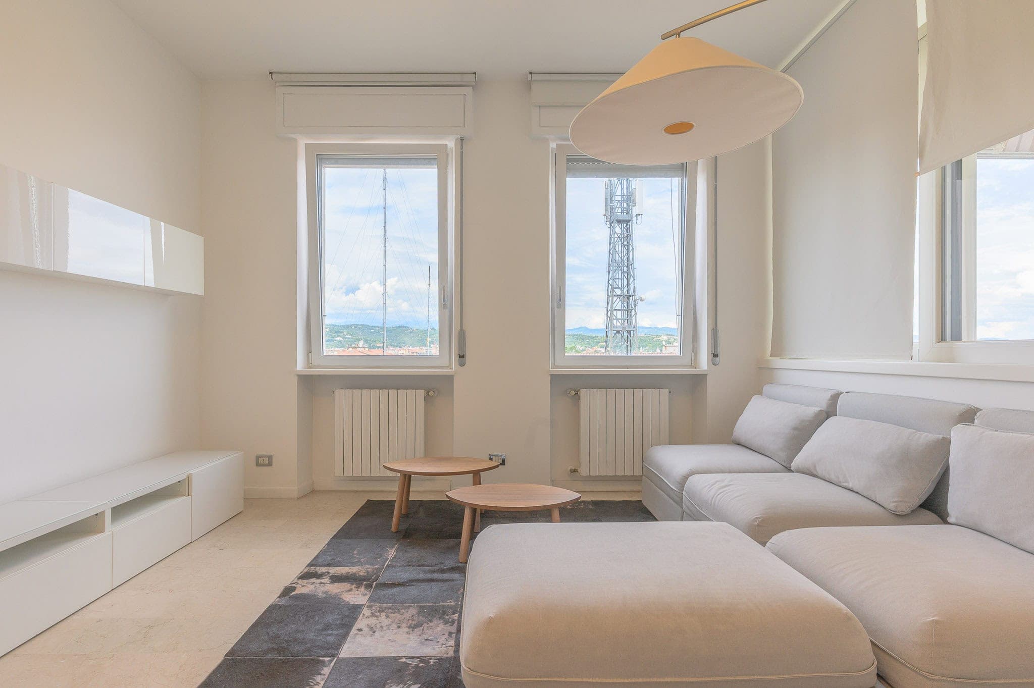 Minimalista soggiorno con grandi finestre, divano bianco e tavolini in legno, vista panoramica su torri e paesaggi verdi, fotografia di Giorgio Zanetti per stile e qualità.