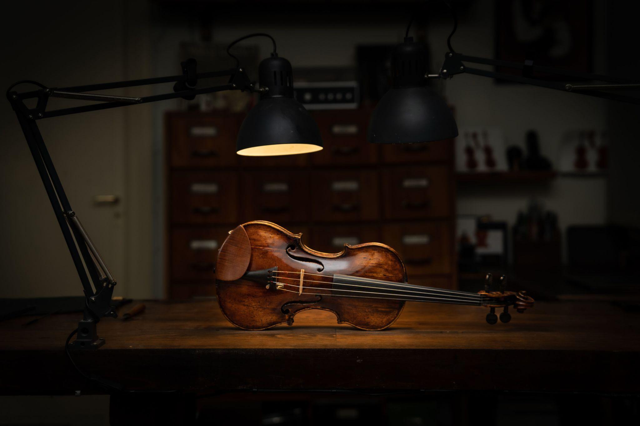 Older man in an apron sits on a stool, holding a violin and talking to a standing young woman in a home music studio with bookshelves behind them.