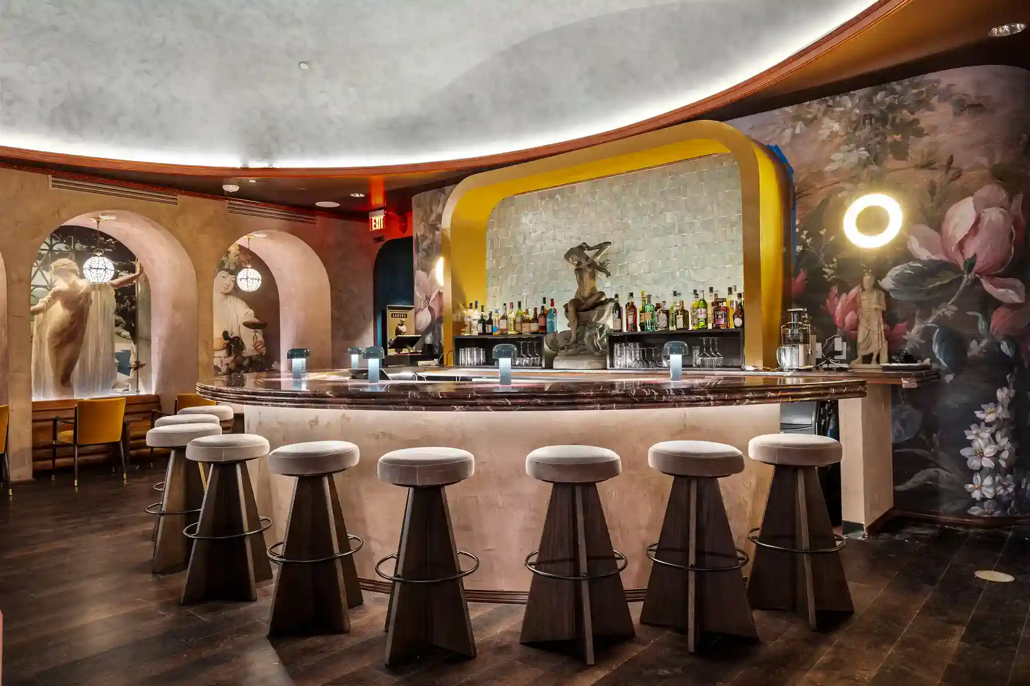 A curved marble bar, upholstered stools, artistic murals, premium liquor display, and warm ambient lighting decorate a luxurious Raleigh bar and cocktail lounge.