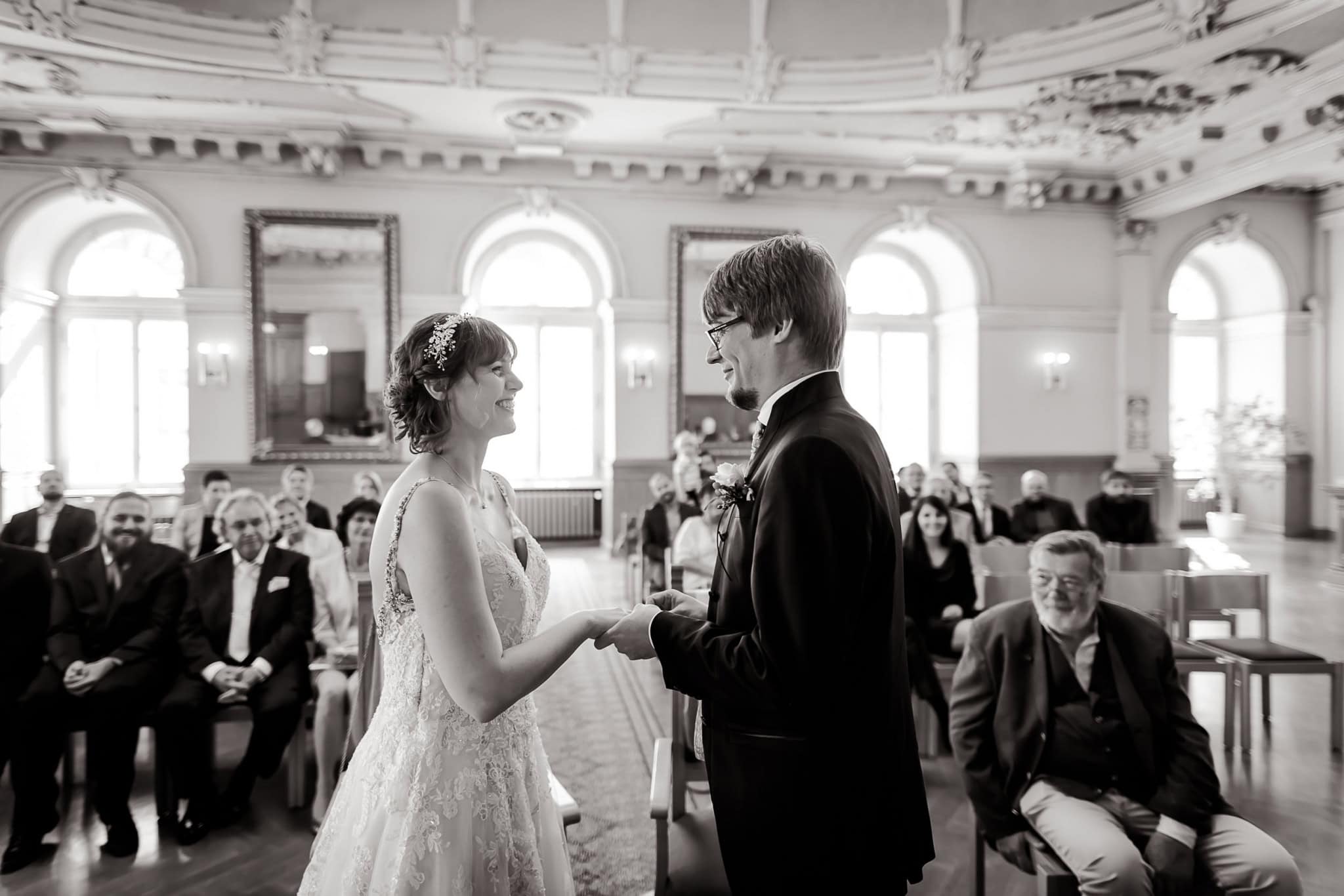 Ein Augenblick inniger Verbundenheit: das Hochzeitspaar hält sich liebevoll die Hände und tauscht verliebte Blicke und die Ringe in einem feierlichen Moment. Umgeben von den Zeugen ihrer Liebe, versprechen sie sich eine gemeinsame Zukunft voller Romantik und Glück