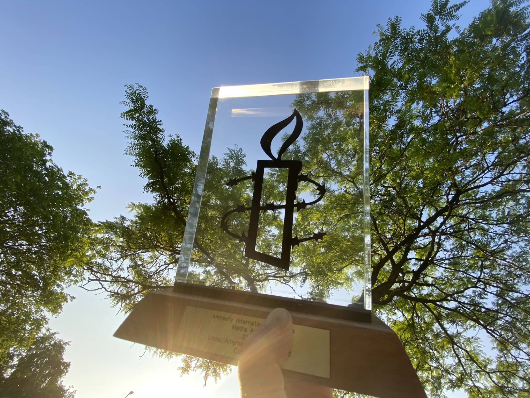 A hand holding a rectangular glass trophy with a brown wooden base under a tree canopy