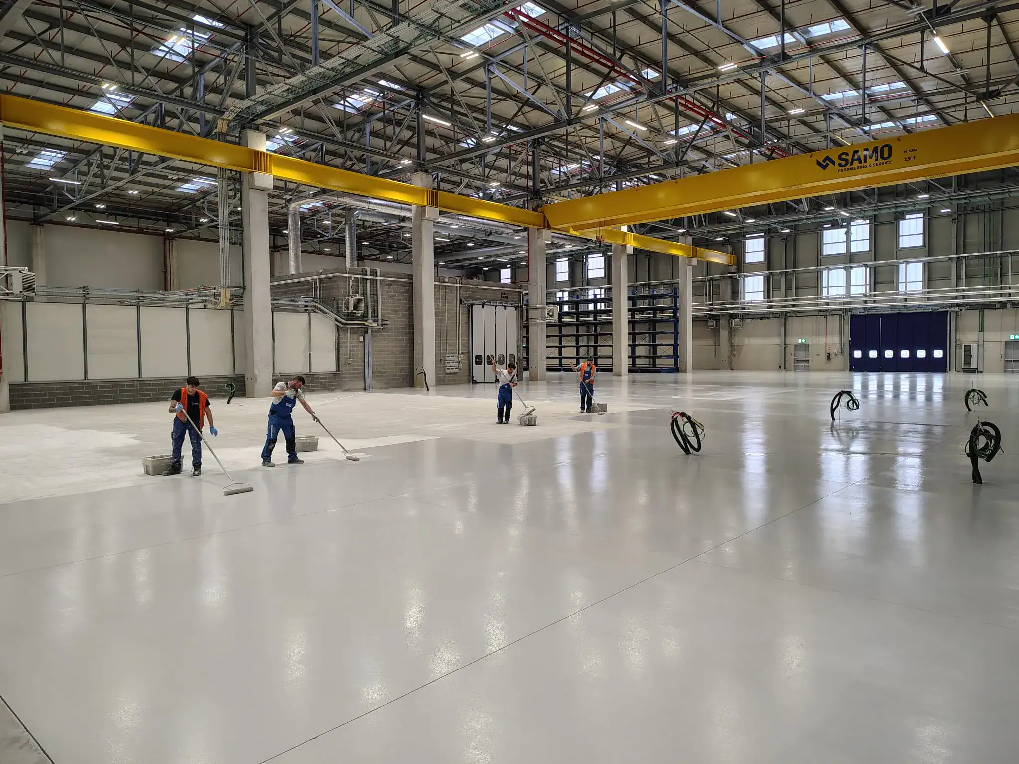 Squadra di applicatori Prima Pavimenti durante la posa di pavimento in resina epossidica nell'hangar dello stabilimento Leonardo Faco di Cameri (NO). Gli operatori in tuta protettiva lavorano sulla superficie in fase di stesura, con i macchinari aeronautici sullo sfondo.