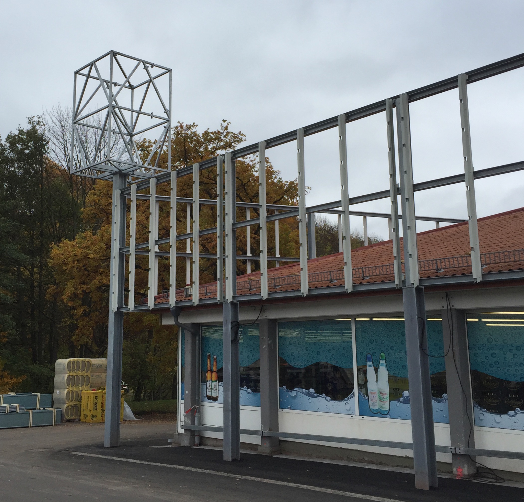 Stahlbau von Stahl- und Metallbau Drost an einem Geschäftsgebäude, mit einem markanten kubischen Stahlgerüst für ein Werbeschild und einer zusätzlichen vertikalen Stahlrahmenkonstruktion entlang der Fassade, vor herbstlichen Bäumen.