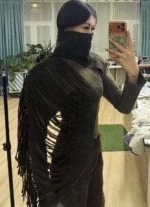 A close-up of a model wearing a dark, felted wool top by Meerim Naizabekova, featuring long, organic fringe and a built-in face covering as part of the "Balance" collection.