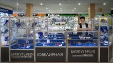 Женщина среднего возраста с короткой стрижкой и открытой улыбкой стоит за стеклянным прилавком-островком. Она в открытой позе, опирается локтями на стеклянную витрину. Одета в светлую блузку и темный жилет. Островок оформлен черными панелями с надписями «БИЖУТЕРИЯ с кристаллами SWAROVSKI» и «ЮВЕЛИРНАЯ». Внутри витрин выставлены украшения (серьги, колье) на синих бархатных подставках. На заднем плане видны другие павильоны ТРЦ, включая вывески «Proficare» и «market».