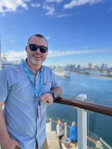 Vacation cruise guest on a balcony overlooking a river and city skyline, enjoying sunny weather and scenic views.