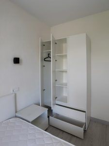 Белый трехстворчатый шкаф с открытыми дверцами, выдвинутыми ящиками и пустой штангой в спальне.