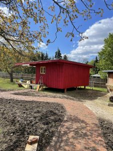 Gemütliches rotes Gartenhaus auf dem Wellnesshof RaJa, umgeben von Natur, mit handgefertigtem Weg und viel Grün. Perfekt für Erholung und Entspannung inmitten idyllischer Umgebung.