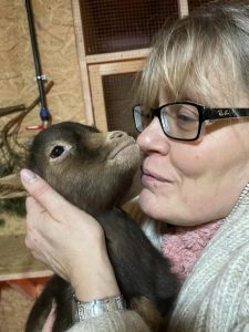 Hochwertiges Bild eines Menschen mit Brille, der ein kleines, braunes Ziegenbaby liebevoll zärtlich hält und küsst, in einer liebevollen und entspannten Atmosphäre im Wellnesshof RaJa.