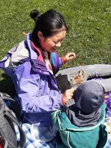 Mom sitting on a picnic mat on grass feeding a chicken wing to a baby facing away from the camera