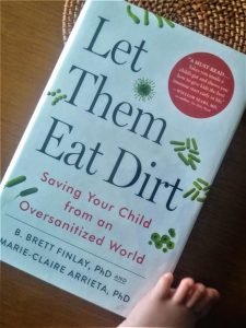 The book Let them Eat Dirt placed on wooden surface with a trivet under it and a babys foot in the bottom right corner