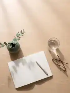 Mesa de madera clara con un cuaderno abierto por una página en blanco, un bolígrafo, un vaso de agua, unas gafas de leer y un jarrón con dos ramas de eucalipto.