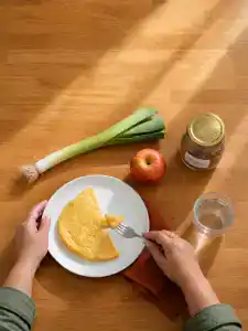 Cenital de una encimera de madera con sol de mañana: unas manos cortan con el tenedor una tortilla francesa en un plato blanco, junto a un puerro, una manzana, un tarro de lentejas y un vaso de agua.