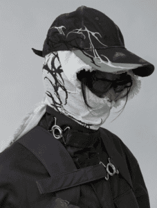 A person wearing a dark cap and sunglasses, with a white FFFPOSTAL scarf featuring black abstract patterns wrapped around their head and neck. They are dressed in a dark jacket with metal rings and straps.