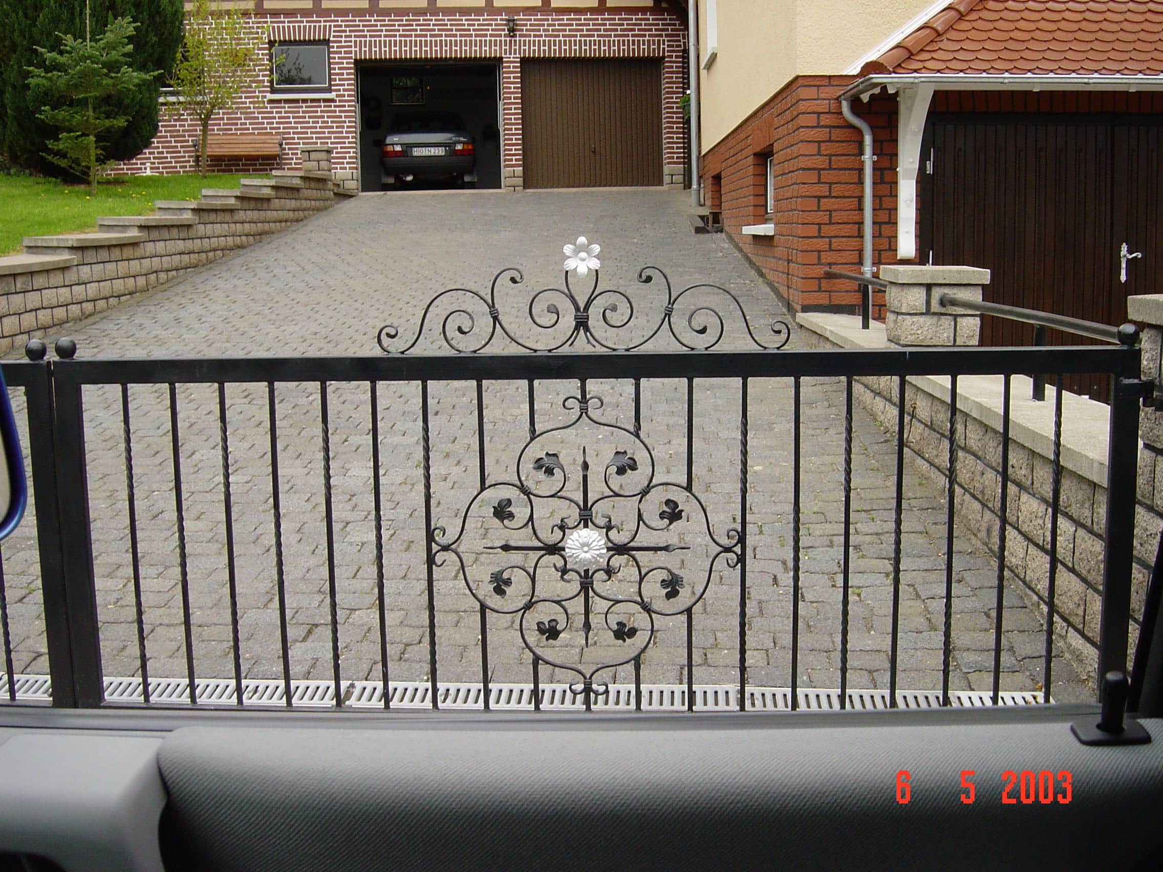 Ein schwarzes, schmiedeeisernes Gartentor von Zaunbau von Stahl- und Metallbau Drost mit kunstvollen Ranken und weißen Blütenmotiven, das eine gepflasterte Auffahrt zu einem Haus mit zwei Garagen abschließt.