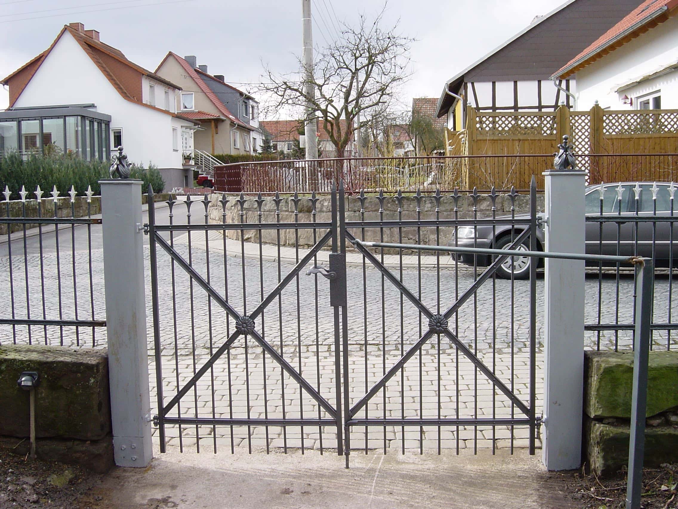 Ein schmiedeeisernes Doppelflügeltor in dunklem Anthrazit, hergestellt von Tore & Torsysteme von Stahl- und Metallbau Drost. Das Tor besitzt vertikale Stäbe, diagonale Kreuzverstrebungen und ist mit kunstvollen floralen Rosetten verziert. Es wird von zwei robusten, hellgrauen Pfosten mit dekorativen Spitzen eingefasst und steht vor einer gepflasterten Einfahrt.