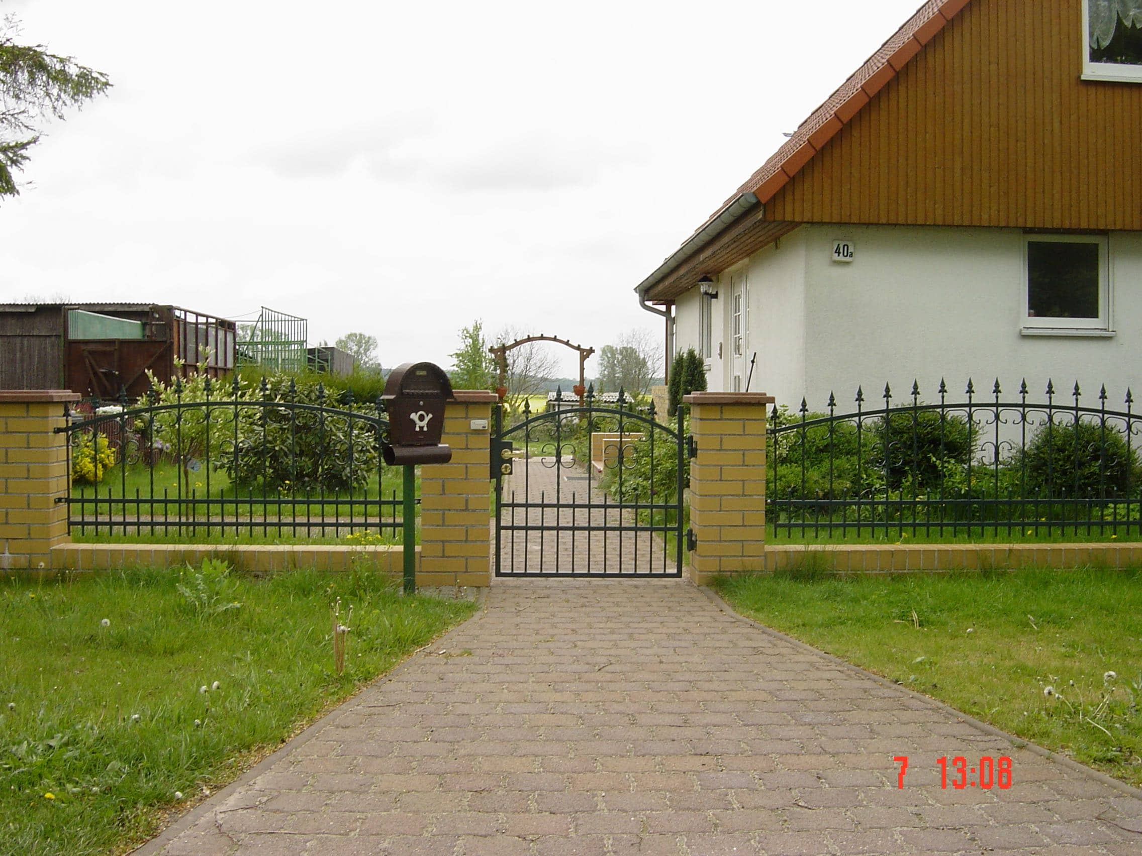 Dunkelgrüner Schmiedeeisenzaun mit elegantem Gartentor und gelben Ziegelpfeilern, eine stilvolle Lösung von Zaunbau von Stahl- und Metallbau Drost vor einem Einfamilienhaus.