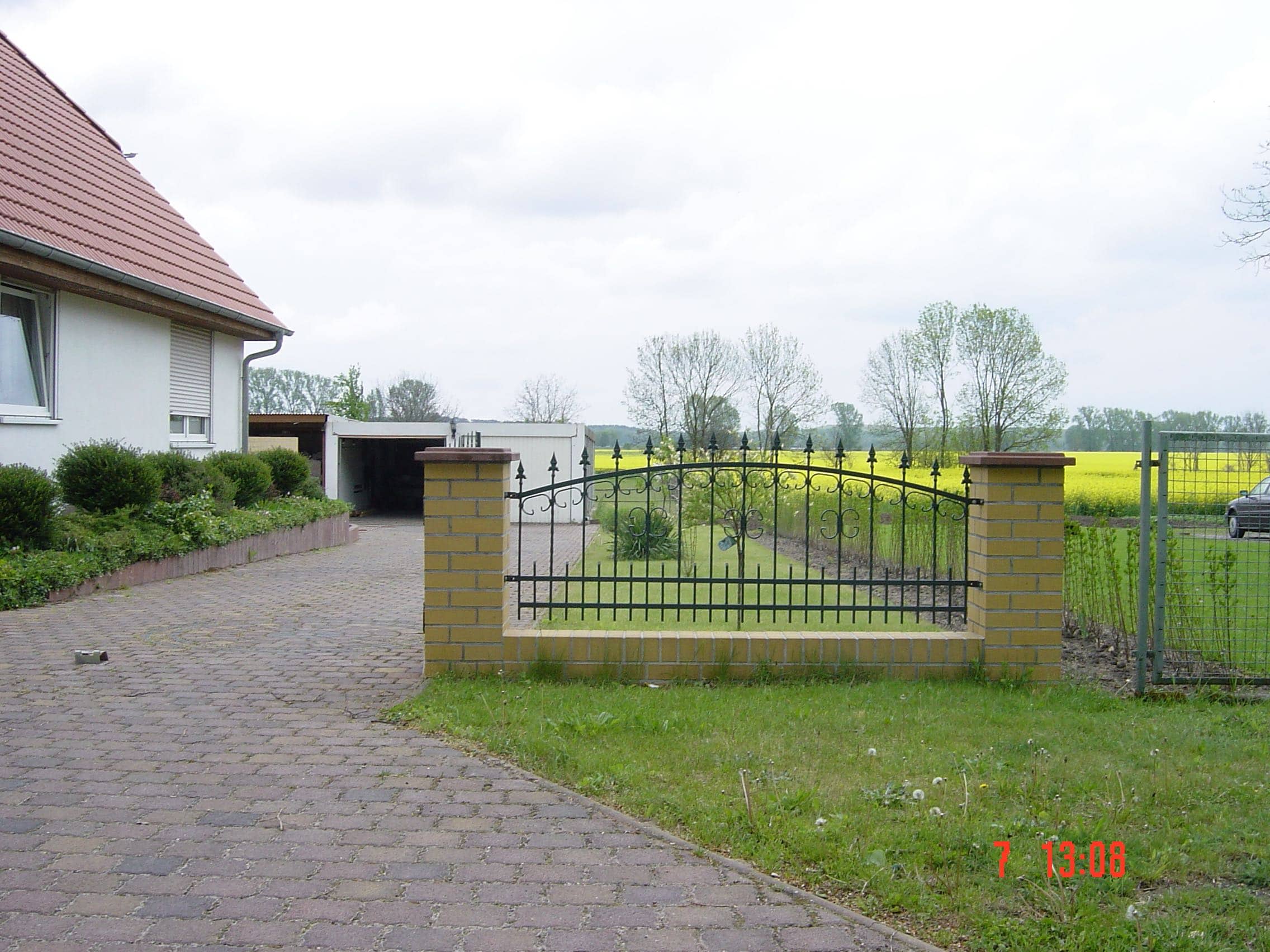 Dekorativer schwarzer Metallzaun mit geschwungenen Elementen und Zierspitzen auf gelben Klinkerpfosten und -sockel, gefertigt von Zaunbau von Stahl- und Metallbau Drost. Der Zaun grenzt einen Vorgarten mit Rasen an eine gepflasterte Einfahrt ab, im Hintergrund sind ein Wohnhaus und ein gelbes Rapsfeld zu erkennen.