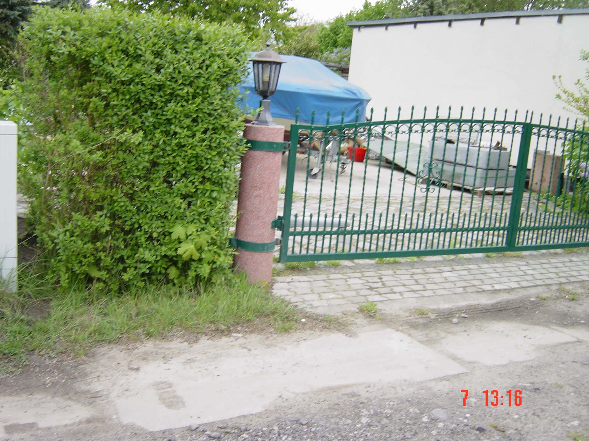Grünes Metalltor mit schmiedeeisernen Spitzen und Zierelementen, befestigt an einem roten Granitpfeiler mit Laterne. Ein Beispiel für Zaunbau von Stahl- und Metallbau Drost.
