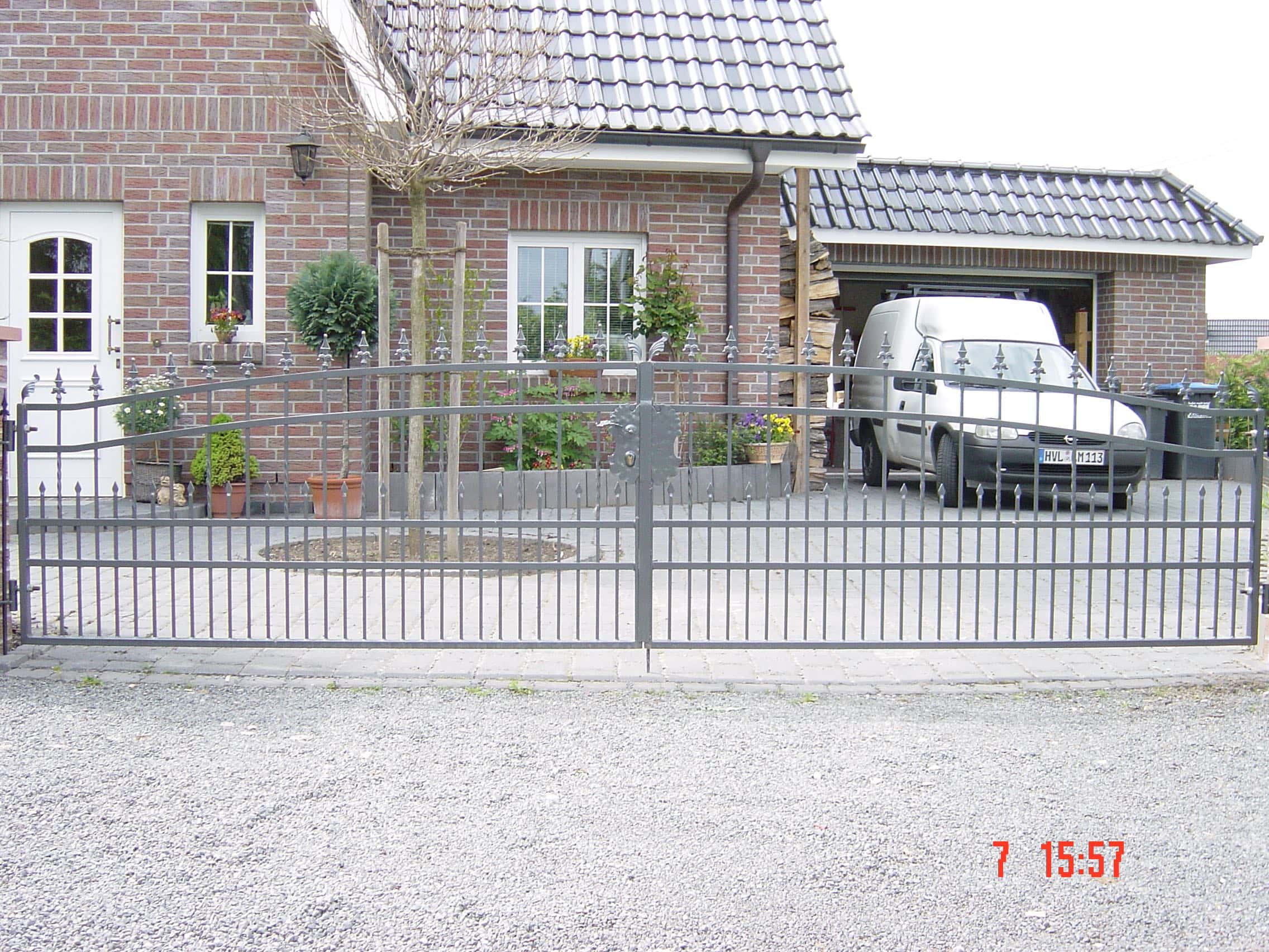Dunkelgraues Metall-Doppelflügeltor mit Zierspitzen und mittigem Schmuckelement, gefertigt von Tore & Torsysteme von Stahl- und Metallbau Drost, vor einem Einfamilienhaus mit Ziegelsteinfassade und einem Lieferwagen im Hintergrund.