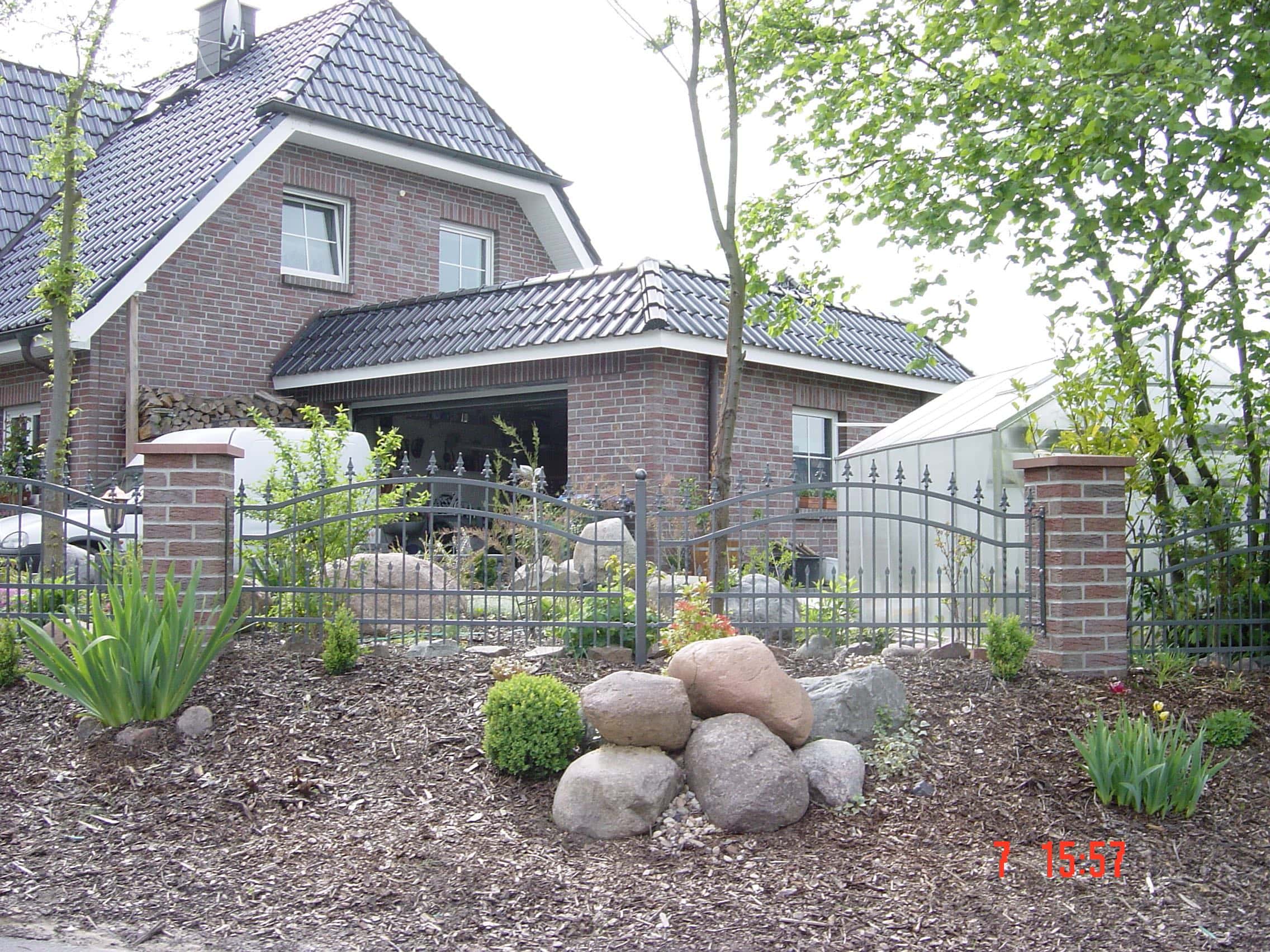 Dunkelgrauer Schmiedezaun von Zaunbau von Stahl- und Metallbau Drost mit gemauerten Pfeilern und dekorativen Elementen vor einem Ziegelhaus mit Gartenanlage.