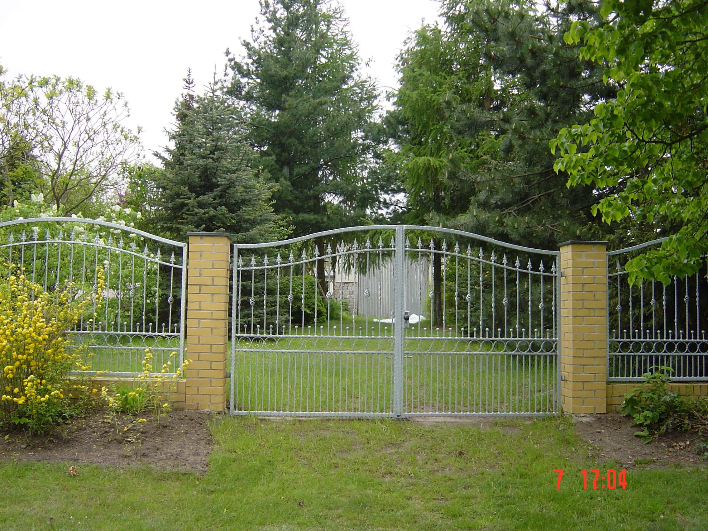 Doppel-Gartentor und Zaun von Zaunbau von Stahl- und Metallbau Drost aus verzinktem Metall mit dekorativen Spitzen und geschwungenen Elementen, flankiert von hellbraunen Ziegelpfeilern, umgeben von grünem Rasen und Bäumen.