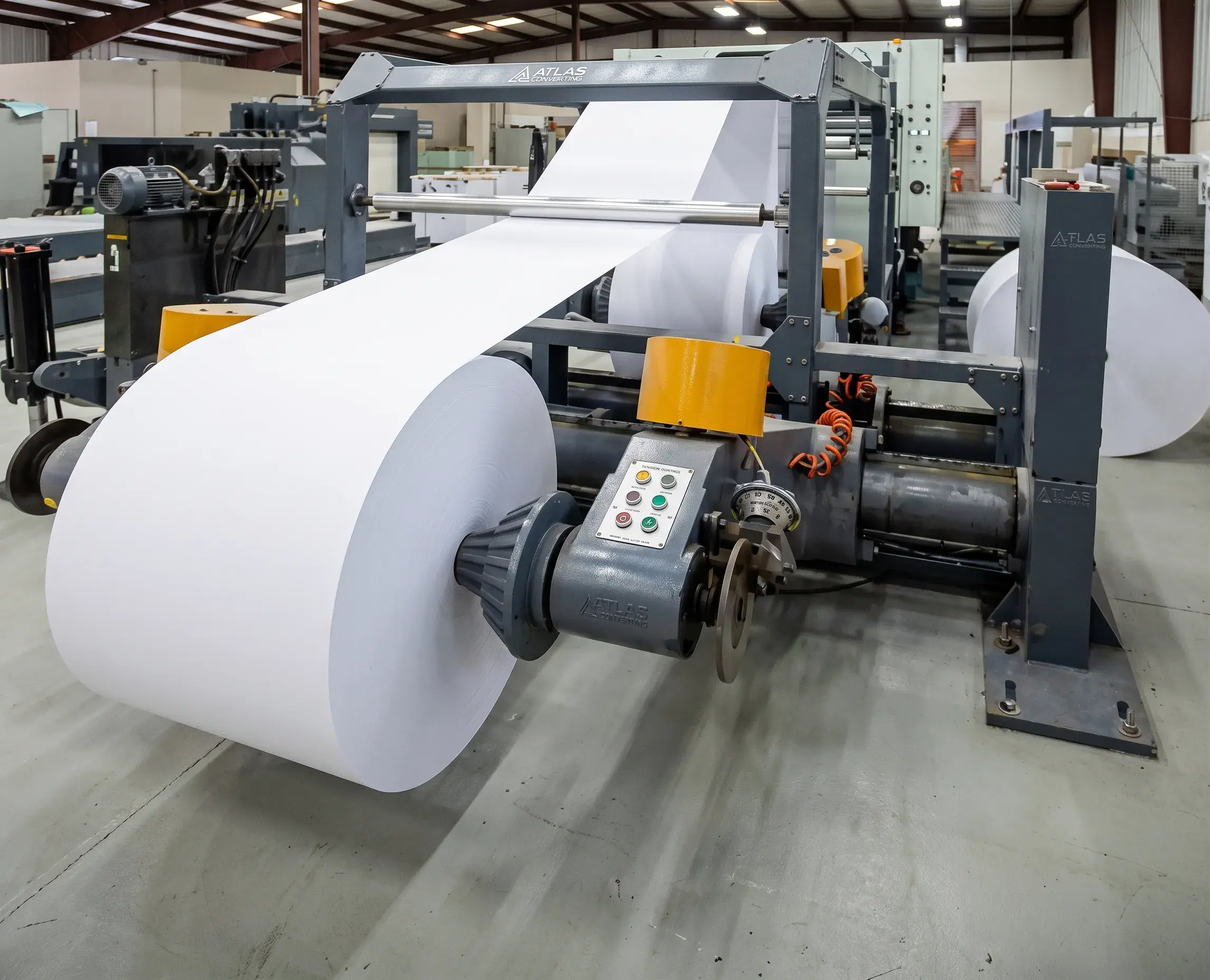 Maquinaria rebobinadora Atlas procesando un gran rollo de papel blanco en una planta industrial.