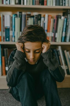 young boy with hands on his head sitting on the floor by books representing the effects of trauma from narcissistic abuse. See a trauma therapist near me today.