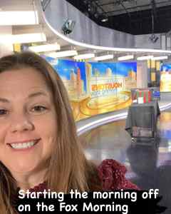 Studio TV set with a woman taking a selfie in the foreground, cityscape graphics on screens, and an ornate stage setup, capturing a behind-the-scenes moment from Fox Morning News.