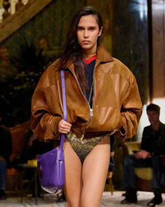 Model in oversized brown leather bomber jacket, metallic briefs, and purple shoulder bag from Versace Spring/Summer 2026 collection.