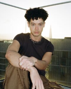A person with curly hair sits on a rooftop at sunset, channeling Erykah Badu vibes in a black t-shirt, brown pants, and a chain necklace, with modern buildings and a glass railing in the background.