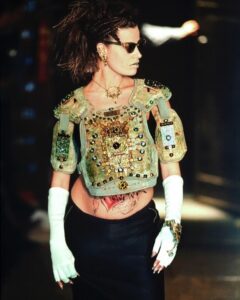 A model wearing sunglasses, a bejeweled gold chest plate, white opera gloves, a long black skirt, and jewelry walks Jean Paul Gaultier 1995 runway, with her hair styled in braids and a voluminous ponytail.