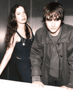 A moody portrait of the Witch Post band members, featuring a woman in a black tank top and a man in a dark trench coat against a metallic backdrop.