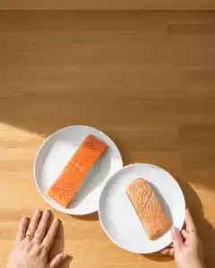 Dos platos blancos sobre una encimera de madera: en uno un lomo de salmón crudo y en el otro el mismo lomo ya cocinado, visiblemente más pequeño, con las manos de una mujer apoyadas al borde de la mesa.