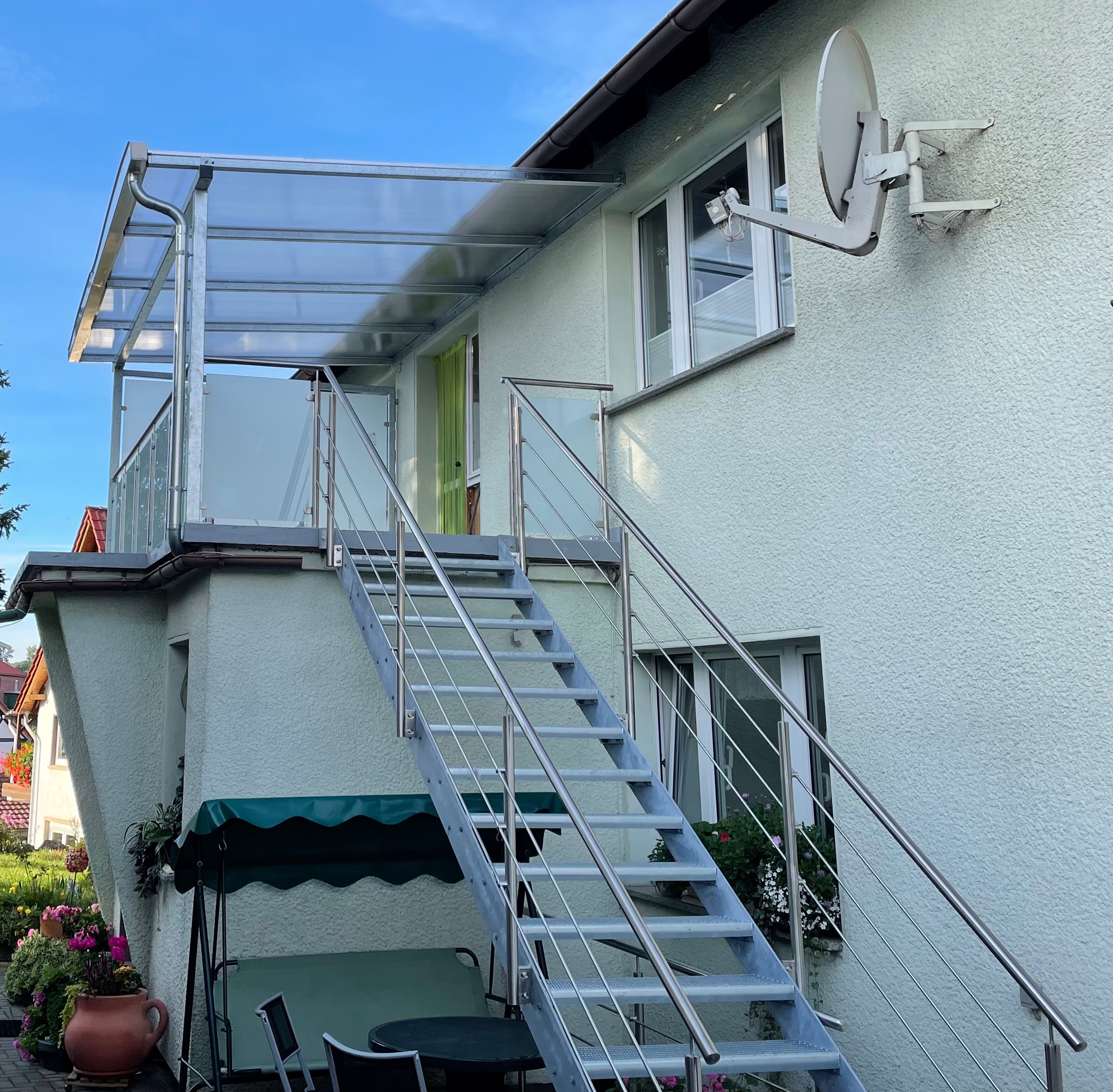 Außenanlage von Stahl- und Metallbau Drost: Eine verzinkte Stahltreppe mit Edelstahlgeländer und Milchglasfüllung führt zu einem überdachten Balkon mit transparentem Dach, an einem hellgrünen Haus.
