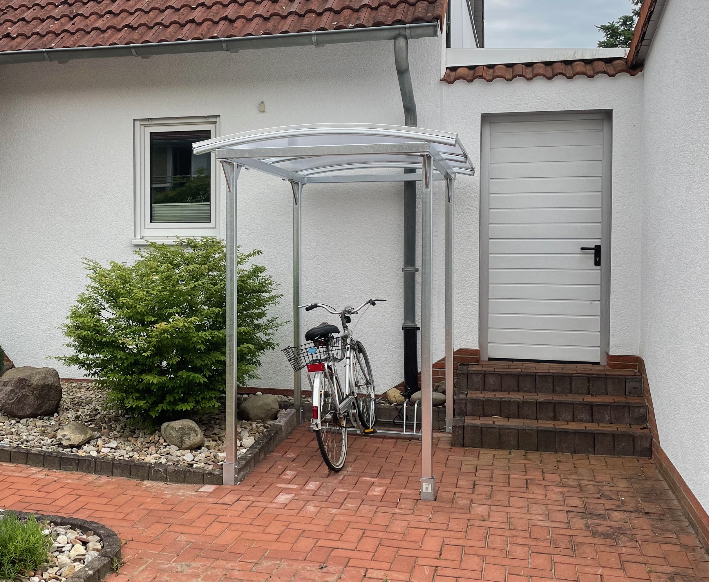 Sonderanfertigungen von Stahl- und Metallbau Drost: Fahrradüberdachung aus verzinktem Stahl mit transparentem Bogendach, schützt ein silbernes Fahrrad vor einem weißen Haus auf Ziegelpflaster.