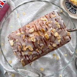 loaf cake topped with icing and toasted almonds on a glass cake stand with a silver slice. Slice of cake on plate to the side.