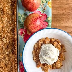 Fresh milled flour apple crisp in a glass pan on top of a brightly colored kitchen placemat. Two apples and a bowl of apple crisp with a scoop of vanilla ice cream is on the side of the glass pan.