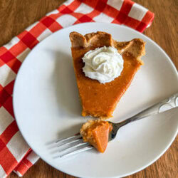 Fresh milled pumpkin pie on top of a white plate with a bite taken out with a fork. White plate is on top of a orange-checkered kitchen towel.