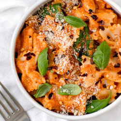 close up of red pepper pasta in a white bowl next to fork