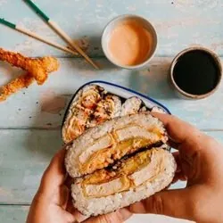 hand holding an onigirazu sandwich, more sandwiches in a bowl, prawn katsu, sauce and chopsticks