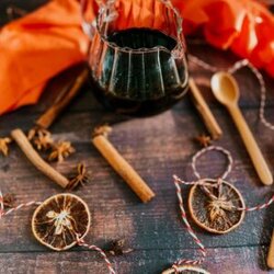 from front - gingerbread syrup in a jug, spoon, star anise, orange slices and cinnamon sticks around, twine, spoons and a red towel on the sides. over a wooden background