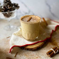 baileys eggnog in a glass, under a red and white towel and wooden coaster, around cinnamon sticks and sugar, in the back pinecones and a marble background