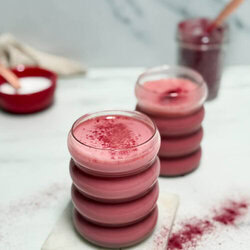 A creamy red latte topped with a dusting of red powder, served in a stackable glass on a white marble coaster. The background features a blurred jar of powder and a red bowl, adding to the cozy vibe.