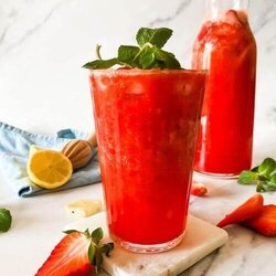 glass of blended strawberry lemonade on a marble coaster, behind a jug of lemonade, strawberries mint and lemons around, in the back a blue towel and lemon squeezer on a marble background